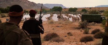 Movie still from “Lord of War” (2005), directed by Andrew Niccol – A man holding a rifle in a field with tents in the background; Extreme Wide shot, Over the shoulder angle