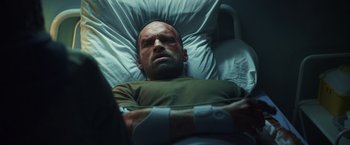 Movie still from “Lost Bullet 2: Back for More” (2022), directed by Guillaume Pierret – A man in a green shirt laying in a hospital bed; Medium shot, Overhead angle