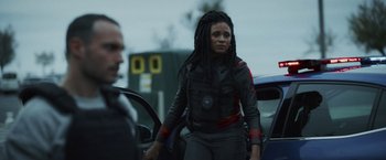 Movie still from “Lost Bullet 2: Back for More” (2022), directed by Guillaume Pierret – A woman with dreadlocks standing next to a car; Medium shot, Over the shoulder angle