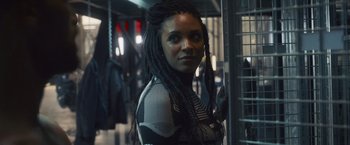 Movie still from “Lost Bullet 2: Back for More” (2022), directed by Guillaume Pierret – A woman with dreads standing next to a metal fence; Close Up shot, Over the shoulder angle