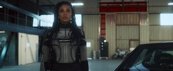 Movie still from “Lost Bullet 2: Back for More” (2022), directed by Guillaume Pierret – A woman with long black dreadlocks standing in a garage; Medium shot, Low angle