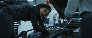 Movie still from “Lost Bullet 2: Back for More” (2022), directed by Guillaume Pierret – A woman looking at the engine of a car in a garage; Medium shot, Low angle