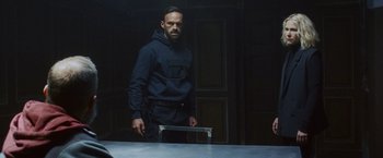 Movie still from “Lost Bullet 2: Back for More” (2022), directed by Guillaume Pierret – A man standing in a dark room holding a metal object; Medium shot, Over the shoulder angle