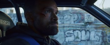 Movie still from “Lost Bullet 2: Back for More” (2022), directed by Guillaume Pierret – A man with a goatee looking out of a car window; Close Up shot, Low angle