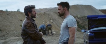 Movie still from “Lost Bullet 2: Back for More” (2022), directed by Guillaume Pierret – Two men are talking in the dirt near a pile of rocks; Medium shot, Over the shoulder angle