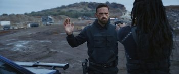 Movie still from “Lost Bullet 2: Back for More” (2022), directed by Guillaume Pierret – A man in a black jacket is holding his hand up to his face; Medium shot, Over the shoulder angle