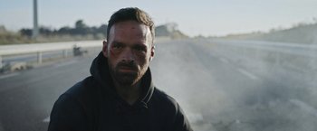 Movie still from “Lost Bullet 2: Back for More” (2022), directed by Guillaume Pierret – A man with a beard is standing in front of a road; Close Up shot, Low angle
