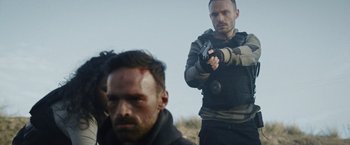 Movie still from “Lost Bullet 2: Back for More” (2022), directed by Guillaume Pierret – A man holding a gun while standing next to another man; Medium shot, Over the shoulder angle