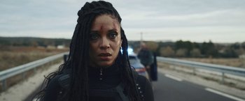 Movie still from “Lost Bullet 2: Back for More” (2022), directed by Guillaume Pierret – A woman with dreadlocks is standing on the side of the road; Close Up shot, Over the shoulder angle