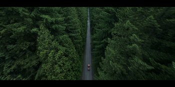 Movie still from “Lou” (2022), directed by Anna Foerster – An aerial view of a car driving down a road through a forest; Extreme Wide shot, Overhead angle