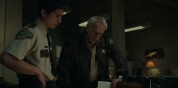 Movie still from “Lou” (2022), directed by Anna Foerster – A man in a police uniform looking at papers with another man; Medium shot, Over the shoulder angle