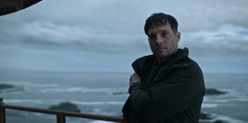 Movie still from “Lou” (2022), directed by Anna Foerster – A man standing on top of a wooden railing near the ocean; Close Up shot, Low angle