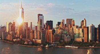Movie still from “Love Again” (2023), directed by Jim Strouse – A view of a city skyline at sunset with a body of water in the foreground; Extreme Wide shot, High angle