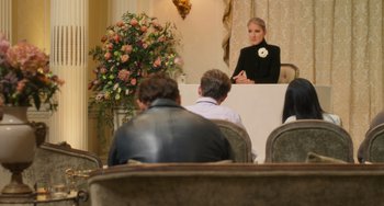 Movie still from “Love Again” (2023), directed by Jim Strouse – A woman sitting at a table in front of a group of people; Wide shot, Over the shoulder angle