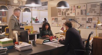 Movie still from “Love Again” (2023), directed by Jim Strouse – Two people sitting at a table in an office setting; Wide shot, High angle
