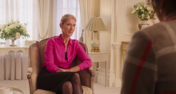 Movie still from “Love Again” (2023), directed by Jim Strouse – A woman in a pink shirt sitting in a chair in a room; Medium shot, Over the shoulder angle
