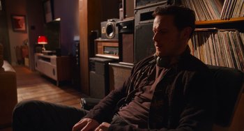 Movie still from “Love Again” (2023), directed by Jim Strouse – A man sitting on a couch in a living room; Medium shot, High angle