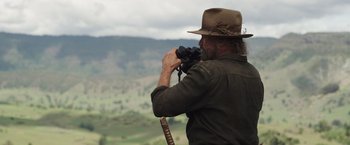Movie still from “Love and Monsters” (2020), directed by Michael Matthews – A man in a hat looking through a pair of binoculars; Medium shot, Over the shoulder angle