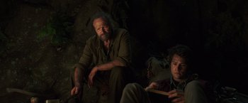 Movie still from “Love and Monsters” (2020), directed by Michael Matthews – A man sitting next to another man in a cave; Medium shot, Low angle