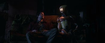 Movie still from “Love and Monsters” (2020), directed by Michael Matthews – A man sitting on a couch next to a robot; Medium shot, Low angle