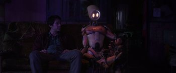 Movie still from “Love and Monsters” (2020), directed by Michael Matthews – A man sitting on a couch next to a robot; Medium shot, Over the shoulder angle