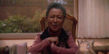 Movie still from “Love Hard” (2021), directed by Hernán Jiménez – An older woman sitting at a table in a wicker chair; Close Up shot, Over the shoulder angle