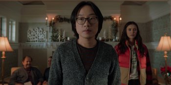 Movie still from “Love Hard” (2021), directed by Hernán Jiménez – A man wearing glasses and a sweater standing in front of other people; Close Up shot, Over the shoulder angle