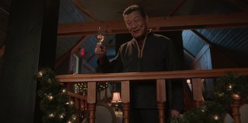 Movie still from “Love Hard” (2021), directed by Hernán Jiménez – An older man standing on a wooden railing holding a candle; Medium shot, Low angle