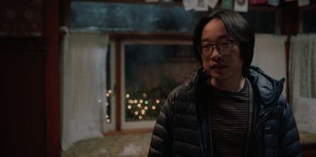 Movie still from “Love Hard” (2021), directed by Hernán Jiménez – A man wearing glasses and a black jacket standing next to a window; Medium shot, Over the shoulder angle