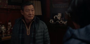 Movie still from “Love Hard” (2021), directed by Hernán Jiménez – An older man wearing a jacket standing in front of a fireplace; Close Up shot, Over the shoulder angle