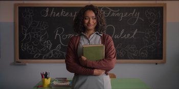 Movie still from “Love in the Villa” (2022), directed by Mark Steven Johnson – A woman standing in front of a chalkboard holding a book; Medium shot, Over the shoulder angle