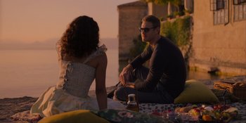 Movie still from “Love in the Villa” (2022), directed by Mark Steven Johnson – A man and a woman sitting on a blanket near the water; Medium shot, Over the shoulder angle