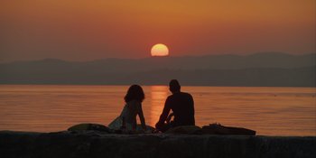 Movie still from “Love in the Villa” (2022), directed by Mark Steven Johnson – A man and a woman sitting on the shore of a body of water at sunset; Extreme Wide shot, High angle