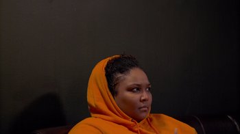 Movie still from “Love, Lizzo” (2022), directed by Doug Pray – A woman wearing an orange hoodie sitting in a chair; Close Up shot, High angle