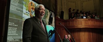 Movie still from “Lucy” (2014), directed by Luc Besson – An older man in a suit speaking at an event; Medium shot, Low angle