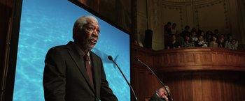 Movie still from “Lucy” (2014), directed by Luc Besson – A man standing at a podium with microphones in front of him; Medium shot, Low angle