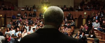 Movie still from “Lucy” (2014), directed by Luc Besson – An older man is standing in front of an audience; Medium shot, Low angle