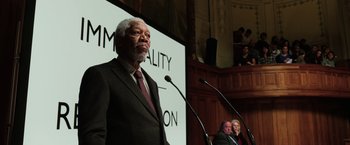 Movie still from “Lucy” (2014), directed by Luc Besson – An older man in a suit and tie speaking at a podium; Medium shot, Low angle