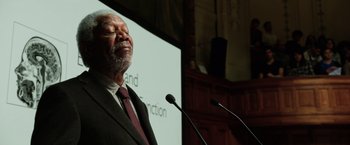 Movie still from “Lucy” (2014), directed by Luc Besson – An older man standing at a podium with microphones in front of him; Close Up shot, Low angle