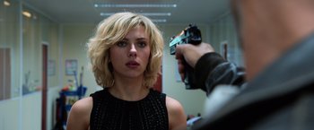 Movie still from “Lucy” (2014), directed by Luc Besson – A man taking a picture of a woman with a gun; Close Up shot, Over the shoulder angle