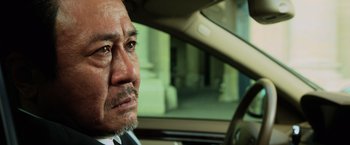 Movie still from “Lucy” (2014), directed by Luc Besson – An older man sitting in the driver's seat of a car; Close Up shot, Over the shoulder angle