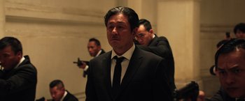 Movie still from “Lucy” (2014), directed by Luc Besson – A man in a suit and tie holding a gun in front of another man in a suit and tie; Medium shot, Low angle