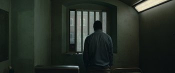 Movie still from “Luther: The Fallen Sun” (2023), directed by Jamie Payne – A man standing in front of a window with bars; Wide shot, Over the shoulder angle