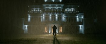 Movie still from “Luther: The Fallen Sun” (2023), directed by Jamie Payne – A person standing in the rain in front of a building; Extreme Wide shot, Low angle