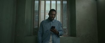 Movie still from “Luther: The Fallen Sun” (2023), directed by Jamie Payne – A man standing in front of a window looking at a cell phone; Medium shot, Low angle