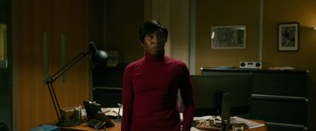 Movie still from “Luther: The Fallen Sun” (2023), directed by Jamie Payne – A man in a red shirt standing in front of a desk; Medium shot, Over the shoulder angle