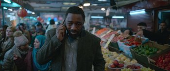 Movie still from “Luther: The Fallen Sun” (2023), directed by Jamie Payne – A man talking on a cell phone while standing in front of a fruit stand; Close Up shot, Over the shoulder angle