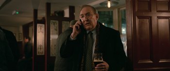 Movie still from “Luther: The Fallen Sun” (2023), directed by Jamie Payne – An older man talking on a cell phone while holding a glass of beer; Close Up shot, Over the shoulder angle