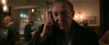 Movie still from “Luther: The Fallen Sun” (2023), directed by Jamie Payne – An older man talking on a cell phone while wearing glasses; Close Up shot, Over the shoulder angle