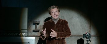 Movie still from “Luther: The Fallen Sun” (2023), directed by Jamie Payne – A man in a brown jacket is talking on a microphone; Medium shot, High angle